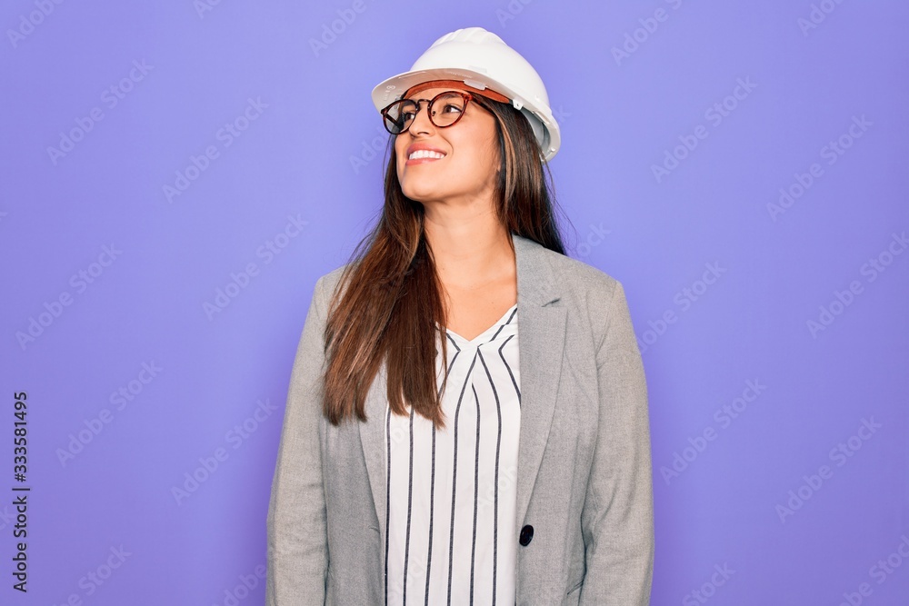 Professional woman engineer wearing industrial safety helmet over pruple background looking away to side with smile on face, natural expression. Laughing confident.