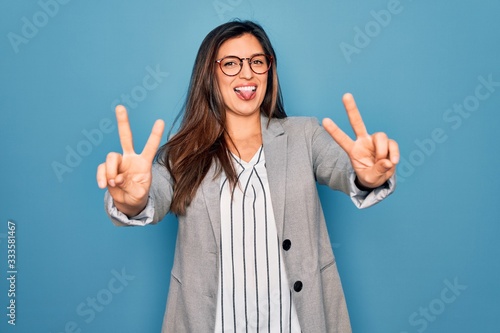 Young hispanic business woman wearing glasses standing over blue isolated background smiling with tongue out showing fingers of both hands doing victory sign. Number two.