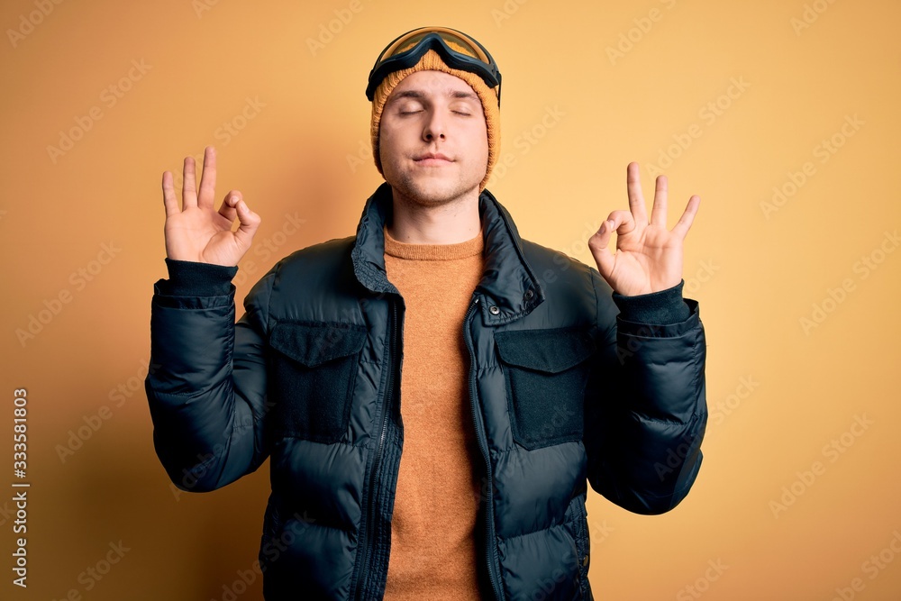 Young handsome caucasian man wearing hat, coat and ski glasses for winter and snow weather relax and smiling with eyes closed doing meditation gesture with fingers. Yoga concept. Young handsome caucasian man wearing hat, coat and ski glasses for winter and snow weather relax and smiling with eyes closed doing meditation gesture with fingers. Yoga concept.