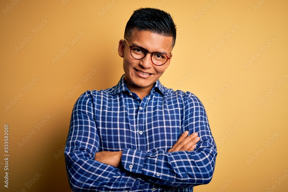 Young handsome latin man wearing casual shirt and glasses over yellow background happy face smiling with crossed arms looking at the camera. Positive person. Young handsome latin man wearing casual shirt and glasses over yellow background happy face smiling with crossed arms looking at the camera. Positive person.