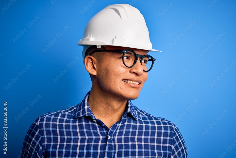 Young handsome engineer latin man wearing safety helmet over isolated blue background looking away to side with smile on face, natural expression. Laughing confident.