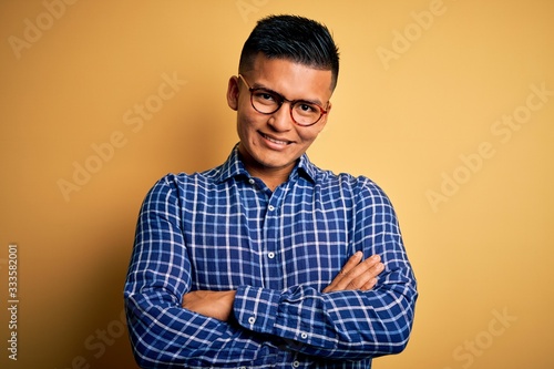 Young handsome latin man wearing casual shirt and glasses over yellow background happy face smiling with crossed arms looking at the camera. Positive person.