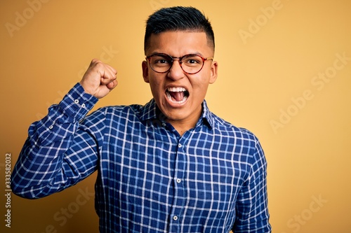 Young handsome latin man wearing casual shirt and glasses over yellow background angry and mad raising fist frustrated and furious while shouting with anger. Rage and aggressive concept.