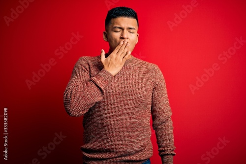 Young handsome latin man wearing casual sweater standing over red background bored yawning tired covering mouth with hand. Restless and sleepiness. © Krakenimages.com