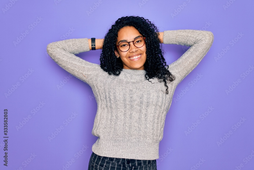Young african american woman wearing casual sweater and glasses over purple background relaxing and stretching, arms and hands behind head and neck smiling happy Young african american woman wearing casual sweater and glasses over purple background relaxing and stretching, arms and hands behind head and neck smiling happy