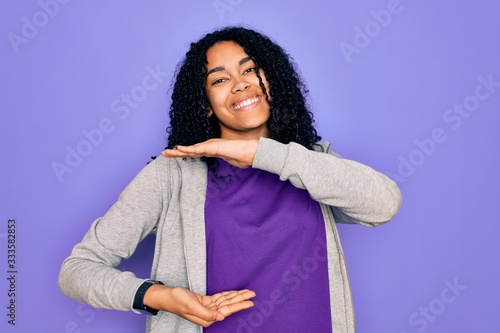 Young african american sporty woman wearing casual sweatshirt over purple background gesturing with hands showing big and large size sign, measure symbol. Smiling looking at the camera. Measuring  © Krakenimages.com