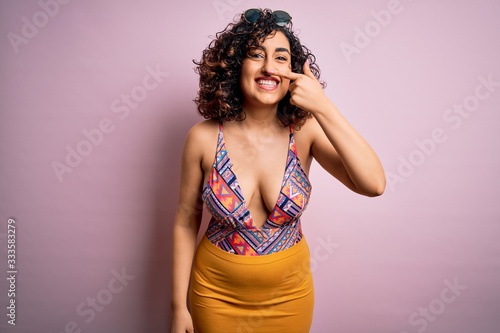 Young beautiful arab woman on vacation wearing swimsuit and sunglasses over pink background Pointing with hand finger to face and nose, smiling cheerful. Beauty concept