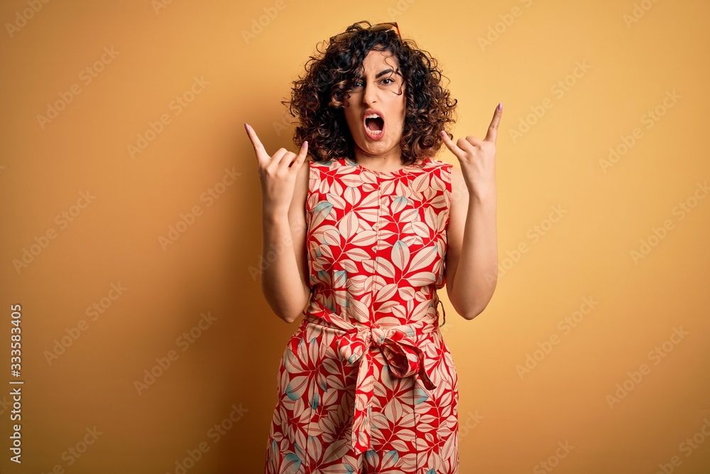 Young beautiful curly arab woman on vacation wearing summer floral dress and sunglasses shouting with crazy expression doing rock symbol with hands up. Music star. Heavy concept. Young beautiful curly arab woman on vacation wearing summer floral dress and sunglasses shouting with crazy expression doing rock symbol with hands up. Music star. Heavy concept.