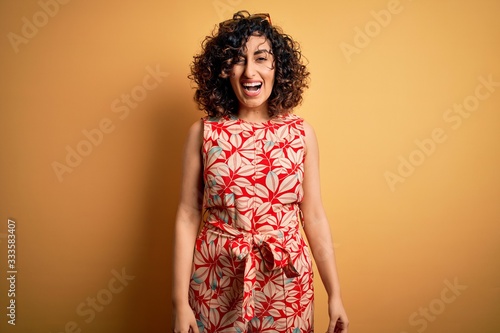 Young beautiful curly arab woman on vacation wearing summer floral dress and sunglasses winking looking at the camera with sexy expression, cheerful and happy face.