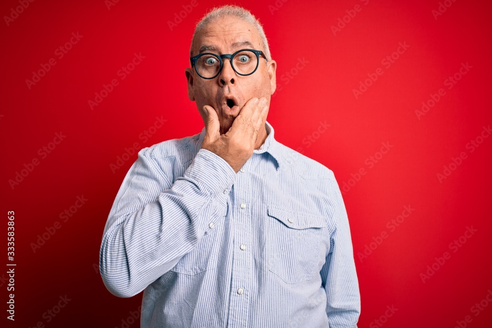 Middle age handsome hoary man wearing casual striped shirt and glasses over red background Looking fascinated with disbelief, surprise and amazed expression with hands on chin