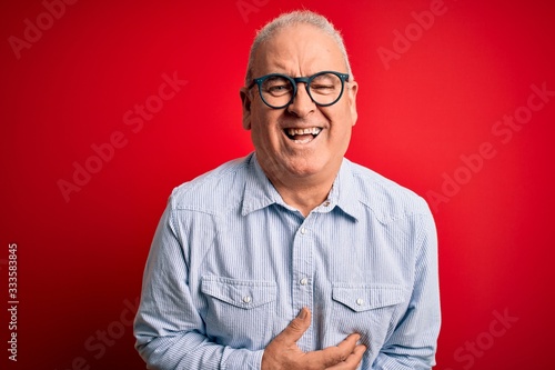 Middle age handsome hoary man wearing casual striped shirt and glasses over red background smiling and laughing hard out loud because funny crazy joke with hands on body. © Krakenimages.com