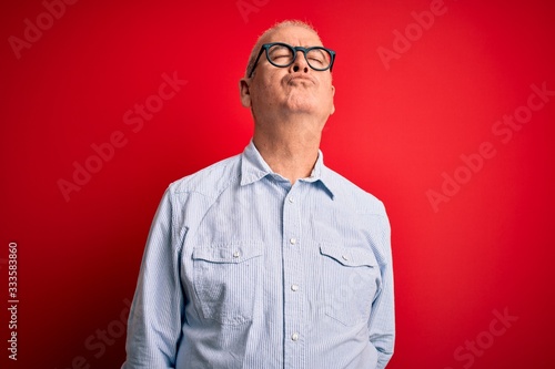 Middle age handsome hoary man wearing casual striped shirt and glasses over red background looking at the camera blowing a kiss on air being lovely and sexy. Love expression.