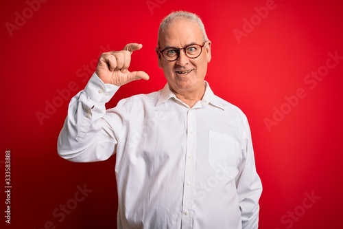 Middle age handsome hoary man wearing casual shirt and glasses over red background smiling and confident gesturing with hand doing small size sign with fingers looking and the camera. Measure concept.