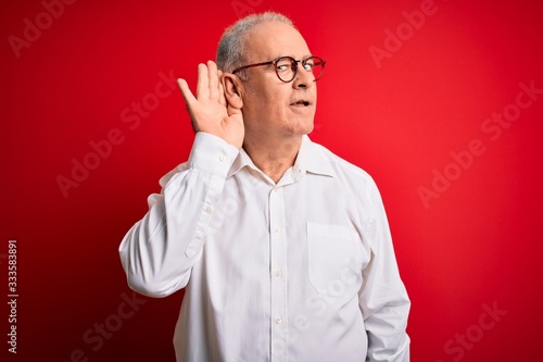 Middle age handsome hoary man wearing casual shirt and glasses over red background smiling with hand over ear listening an hearing to rumor or gossip. Deafness concept. © Krakenimages.com