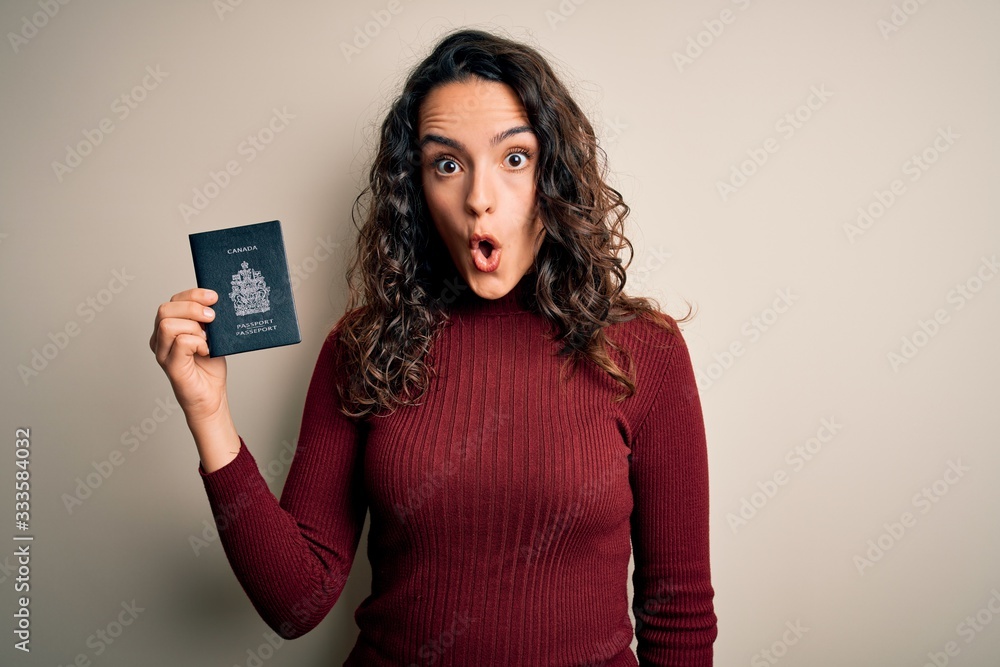Young beautiful tourist woman with curly hair on vacation holding canadian canada passport scared in shock with a surprise face, afraid and excited with fear expression Young beautiful tourist woman with curly hair on vacation holding canadian canada passport scared in shock with a surprise face, afraid and excited with fear expression
