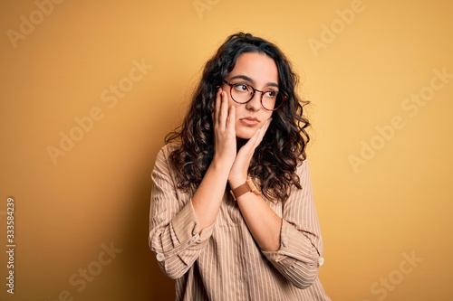 Beautiful woman with curly hair wearing striped shirt and glasses over yellow background Tired hands covering face, depression and sadness, upset and irritated for problem