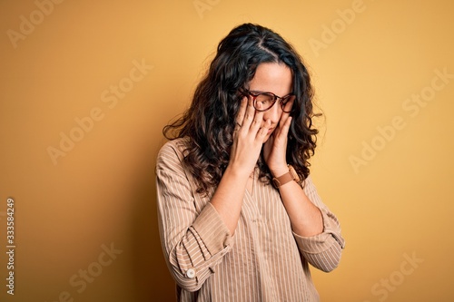 Photos Beautiful woman with curly hair wearing striped shirt and glasses over yellow background rubbing eyes for fatigue and headache, sleepy and tired expression