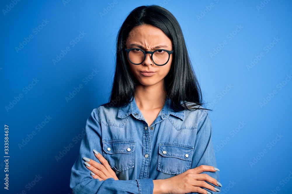 Young beautiful chinese woman wearing casual denim shirt over isolated blue background skeptic and nervous, disapproving expression on face with crossed arms. Negative person. Young beautiful chinese woman wearing casual denim shirt over isolated blue background skeptic and nervous, disapproving expression on face with crossed arms. Negative person.