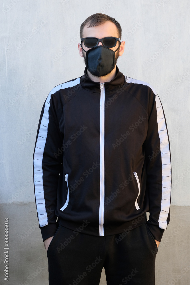 A guy in black glasses and a black face mask is standing against a gray wall. Black cotton masks for protection against bacteria and the spread of the virus. Brunette guy in sportswear hands in pocket A guy in black glasses and a black face mask is standing against a gray wall. Black cotton masks for protection against bacteria and the spread of the virus. Brunette guy in sportswear hands in pocket