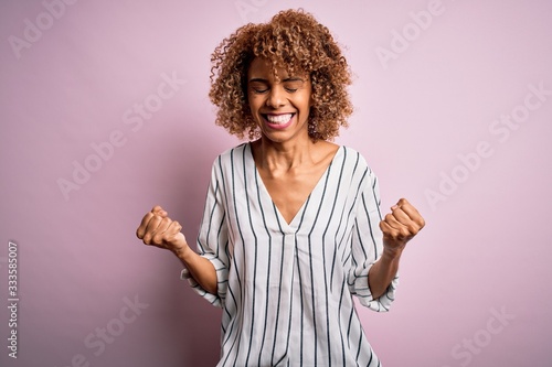 Beautiful african american woman with curly hair wearing striped t-shirt over pink background very happy and excited doing winner gesture with arms raised, smiling and screaming for success. Celebrate