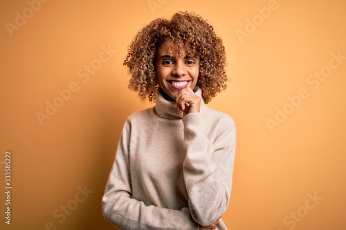Young beautiful african american woman wearing turtleneck sweater over yellow background looking confident at the camera smiling with crossed arms and hand raised on chin. Thinking positive.