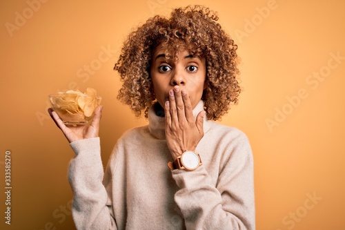 Young african american curly woman holding bowl with chips potatoes over yellow background cover mouth with hand shocked with shame for mistake, expression of fear, scared in silence, secret concept