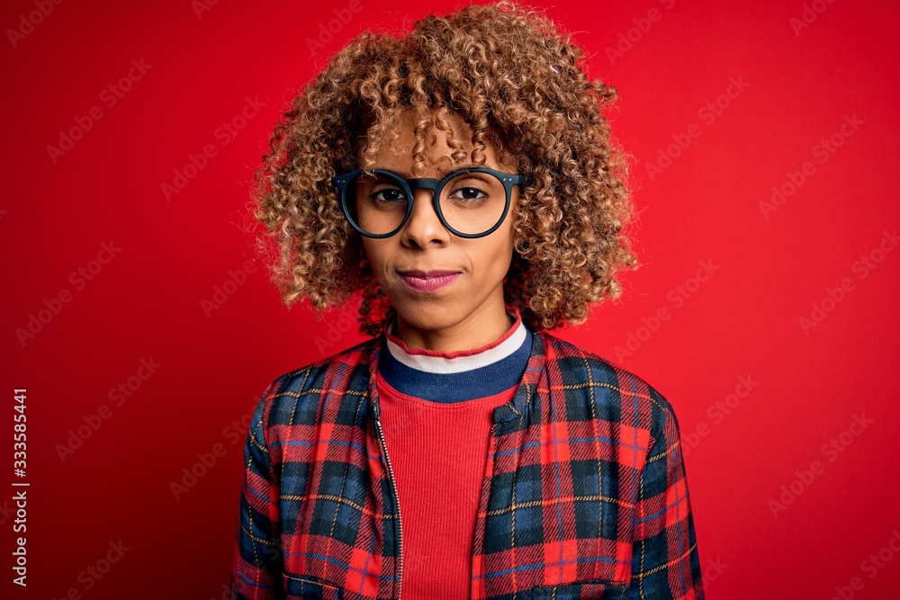Young beautiful african american woman wearing casual shirt and glasses over red background with serious expression on face. Simple and natural looking at the camera.