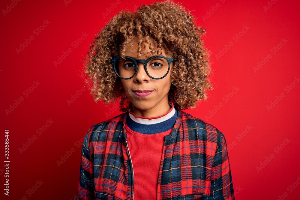 Young beautiful african american woman wearing casual shirt and glasses over red background with serious expression on face. Simple and natural looking at the camera. Young beautiful african american woman wearing casual shirt and glasses over red background with serious expression on face. Simple and natural looking at the camera.