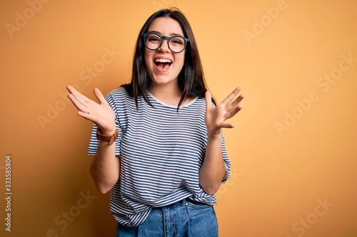 Young brunette woman wearing glasses and navy t-shirt over yellow isolated background celebrating crazy and amazed for success with arms raised and open eyes screaming excited. Winner concept