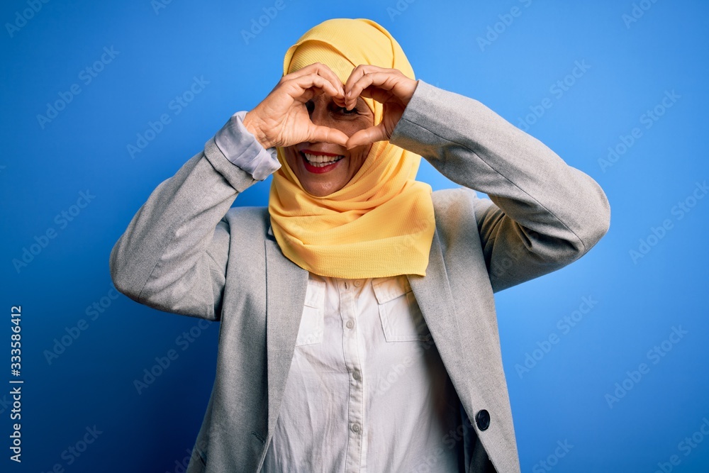 Naklejka premium Middle age brunette business woman wearing muslim traditional hijab over blue background Doing heart shape with hand and fingers smiling looking through sign