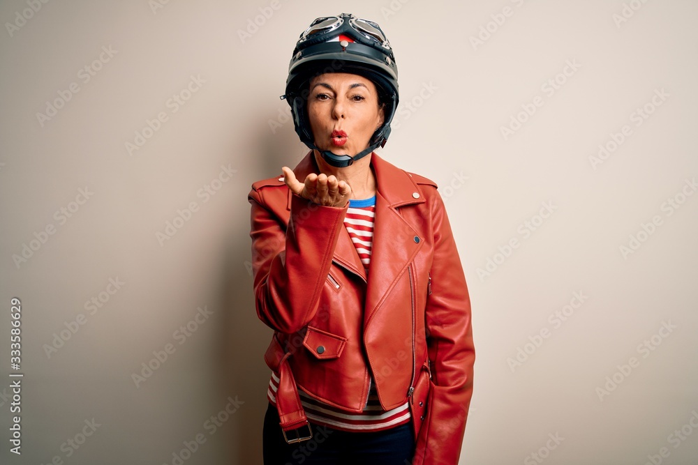 Middle age motorcyclist woman wearing motorcycle helmet and jacket over white background looking at the camera blowing a kiss with hand on air being lovely and sexy. Love expression. Middle age motorcyclist woman wearing motorcycle helmet and jacket over white background looking at the camera blowing a kiss with hand on air being lovely and sexy. Love expression.