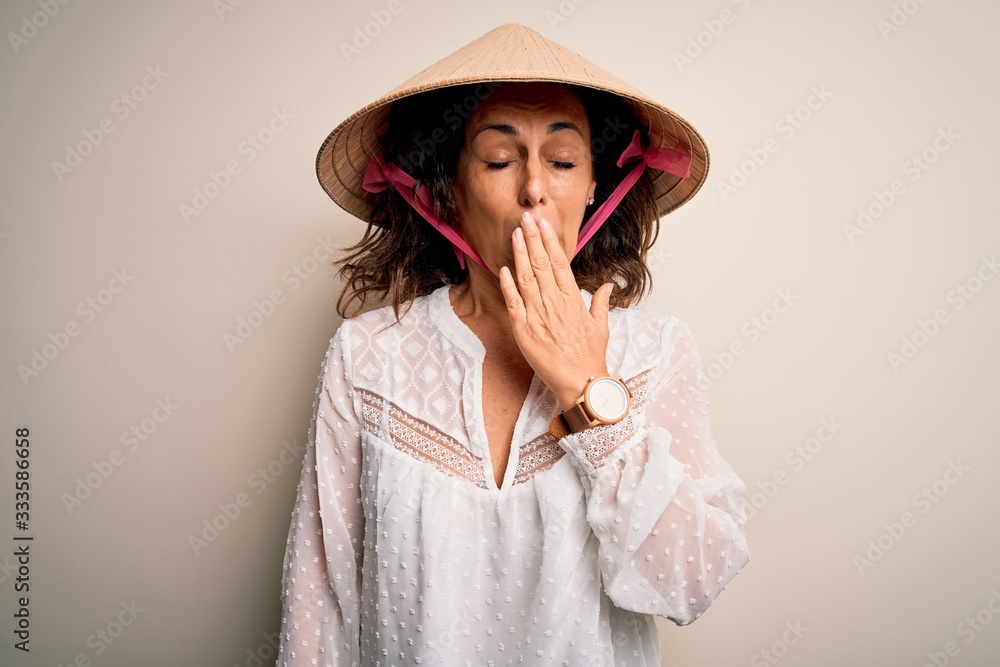 Middle age brunette woman wearing asian traditional conical hat over white background bored yawning tired covering mouth with hand. Restless and sleepiness. Middle age brunette woman wearing asian traditional conical hat over white background bored yawning tired covering mouth with hand. Restless and sleepiness.