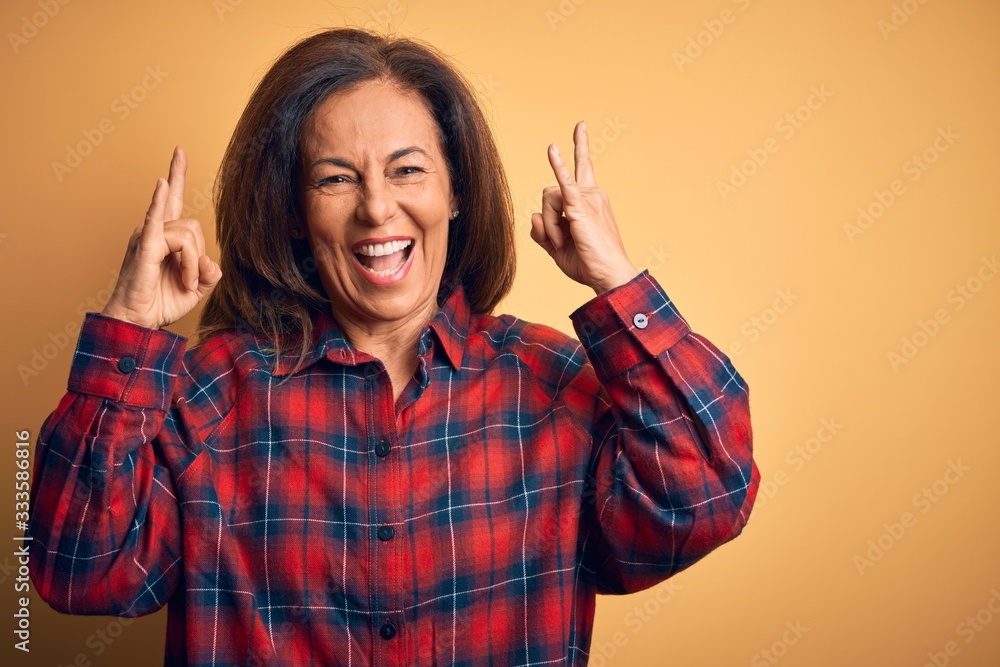 Middle age beautiful woman wearing casual shirt standing over isolated yellow background shouting with crazy expression doing rock symbol with hands up. Music star. Heavy music concept. Middle age beautiful woman wearing casual shirt standing over isolated yellow background shouting with crazy expression doing rock symbol with hands up. Music star. Heavy music concept.