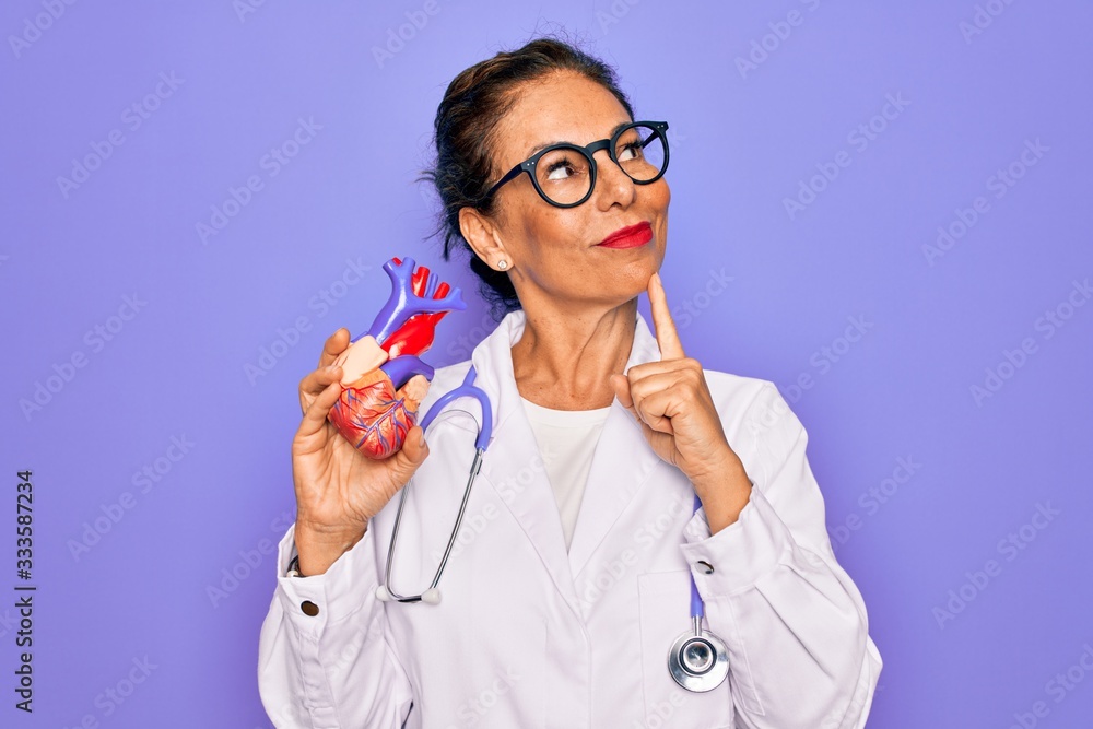 Middle age senior cardiologist doctor woman holding professional cardiology heart serious face thinking about question, very confused idea Middle age senior cardiologist doctor woman holding professional cardiology heart serious face thinking about question, very confused idea