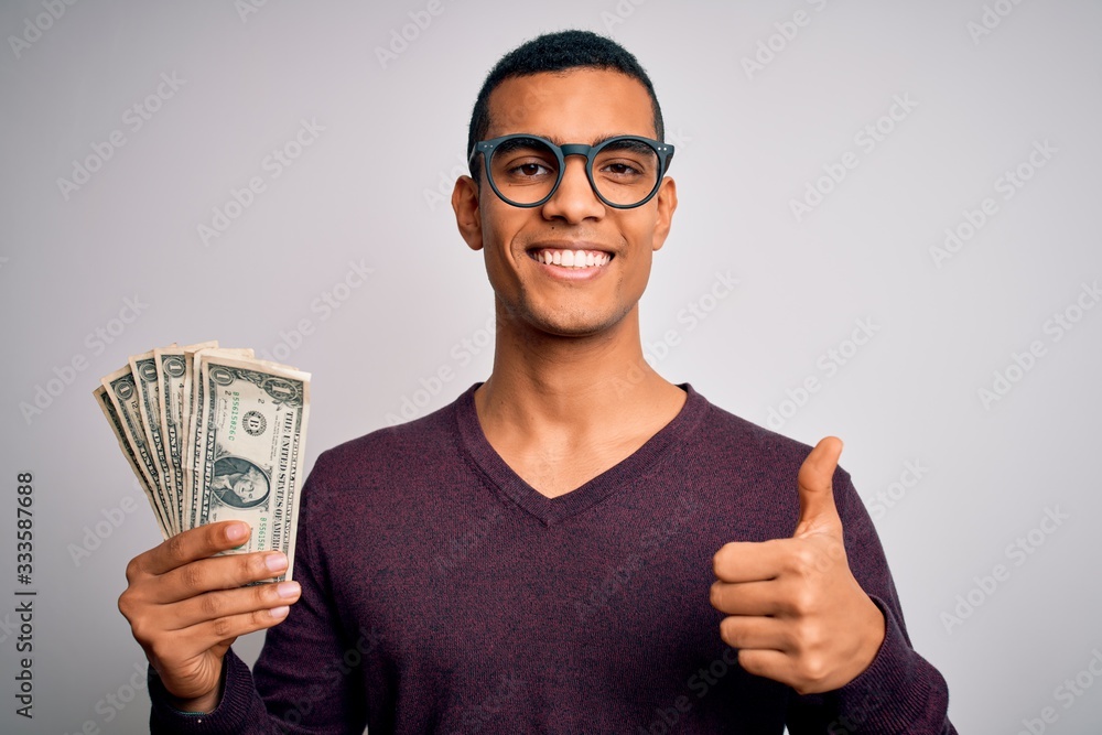 Handsome african american man holding bunch of dollars banknotes over white background happy with big smile doing ok sign, thumb up with fingers, excellent sign Handsome african american man holding bunch of dollars banknotes over white background happy with big smile doing ok sign, thumb up with fingers, excellent sign