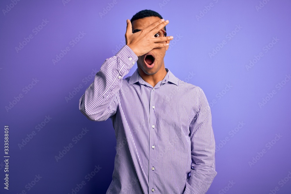 Handsome african american man wearing striped shirt and glasses over purple background peeking in shock covering face and eyes with hand, looking through fingers with embarrassed expression. Handsome african american man wearing striped shirt and glasses over purple background peeking in shock covering face and eyes with hand, looking through fingers with embarrassed expression.