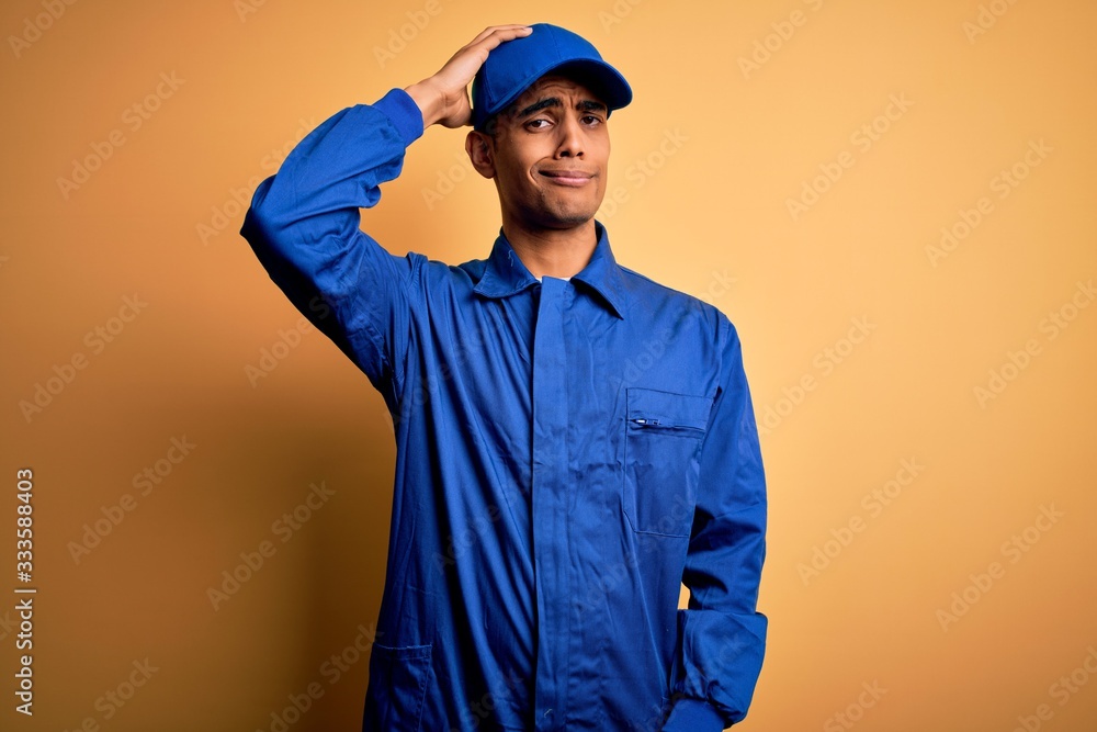 Young african american mechanic man wearing blue uniform and cap over yellow background confuse and wonder about question. Uncertain with doubt, thinking with hand on head. Pensive concept.