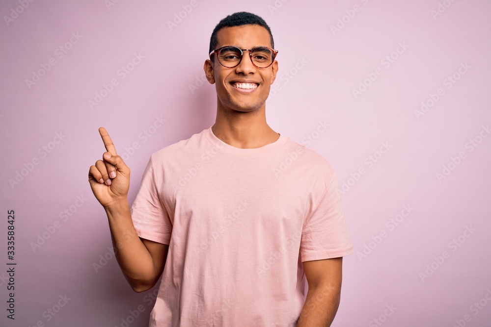 Handsome african american man wearing casual t-shirt and glasses over pink background with a big smile on face, pointing with hand finger to the side looking at the camera. Handsome african american man wearing casual t-shirt and glasses over pink background with a big smile on face, pointing with hand finger to the side looking at the camera.