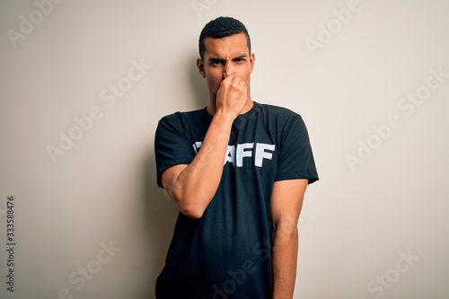 Young handsome african american worker man wearing staff uniform over white background smelling something stinky and disgusting, intolerable smell, holding breath with fingers on nose. Bad smell