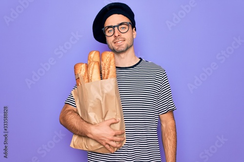 Young handsome baker man with blue eyes wearing french beret holding bag with bread with a happy face standing and smiling with a confident smile showing teeth photo