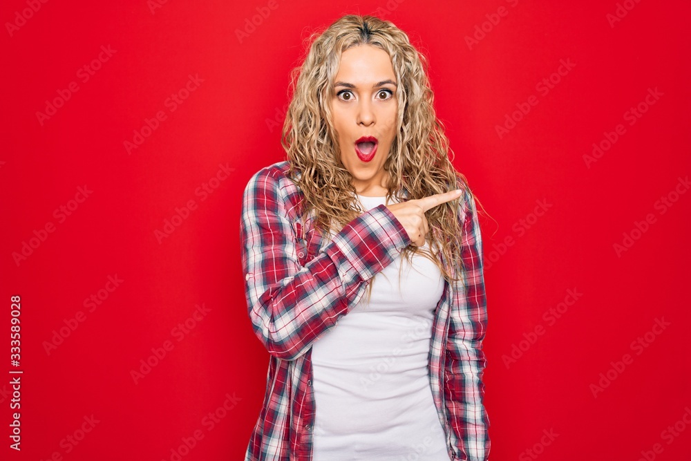Young beautiful blonde woman wearing casual shirt standing over isolated red background Surprised pointing with finger to the side, open mouth amazed expression. Young beautiful blonde woman wearing casual shirt standing over isolated red background Surprised pointing with finger to the side, open mouth amazed expression.