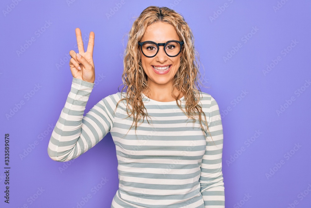 Beautiful blonde woman wearing casual striped t-shirt and glasses over purple background showing and pointing up with fingers number two while smiling confident and happy. Beautiful blonde woman wearing casual striped t-shirt and glasses over purple background showing and pointing up with fingers number two while smiling confident and happy.