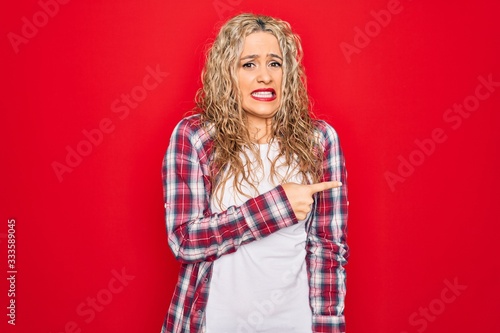 Young beautiful blonde woman wearing casual shirt standing over isolated red background Pointing aside worried and nervous with forefinger, concerned and surprised expression