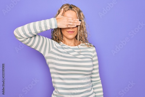 Beautiful blonde woman wearing casual striped t-shirt and glasses over purple background covering eyes with hand, looking serious and sad. Sightless, hiding and rejection concept