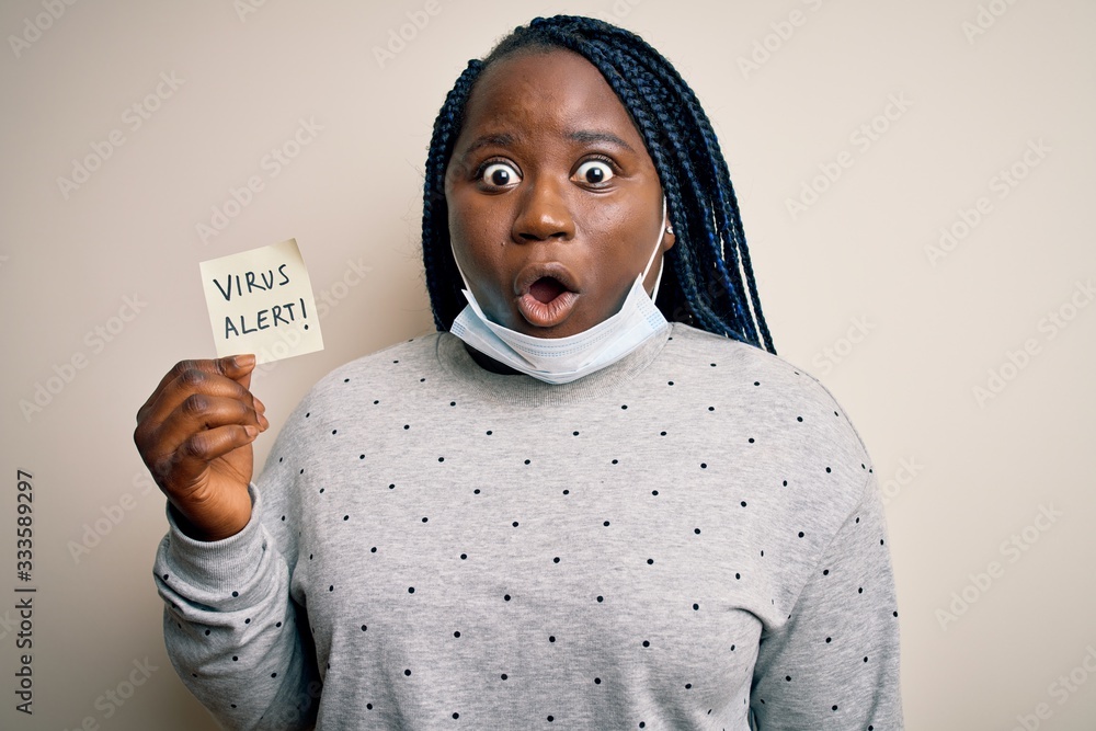 African american woman wearing medical mask holding reminder with virus alert message scared in shock with a surprise face, afraid and excited with fear expression African american woman wearing medical mask holding reminder with virus alert message scared in shock with a surprise face, afraid and excited with fear expression