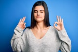 Young beautiful brunette woman wearing casual sweater standing over blue background relax and smiling with eyes closed doing meditation gesture with fingers. Yoga concept.