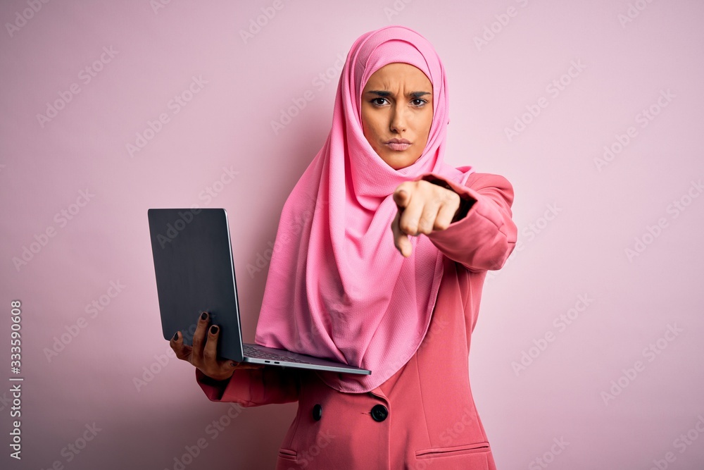 Young beautiful businesswoman wearing pink muslim hijab and business jacket using laptop pointing with finger to the camera and to you, hand sign, positive and confident gesture from the front Young beautiful businesswoman wearing pink muslim hijab and business jacket using laptop pointing with finger to the camera and to you, hand sign, positive and confident gesture from the front