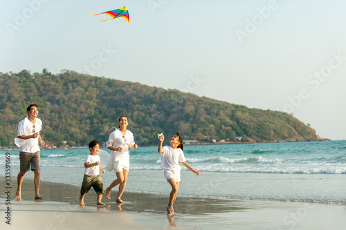 Young Asian happy family parents with child running and having fun with playing kite together on the beach in summertime. Father, mother and kids relax and enjoy summer lifestyle travel vacation
