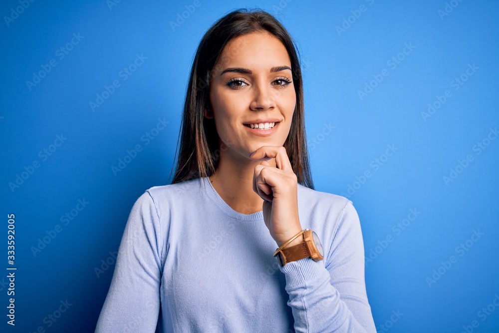 Young beautiful brunette woman wearing casual sweater standing over blue background looking confident at the camera smiling with crossed arms and hand raised on chin. Thinking positive. Young beautiful brunette woman wearing casual sweater standing over blue background looking confident at the camera smiling with crossed arms and hand raised on chin. Thinking positive.