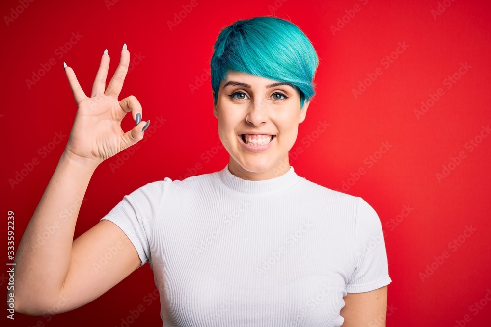 Young beautiful woman with blue fashion hair wearing casual t-shirt over red background smiling positive doing ok sign with hand and fingers. Successful expression. Young beautiful woman with blue fashion hair wearing casual t-shirt over red background smiling positive doing ok sign with hand and fingers. Successful expression.